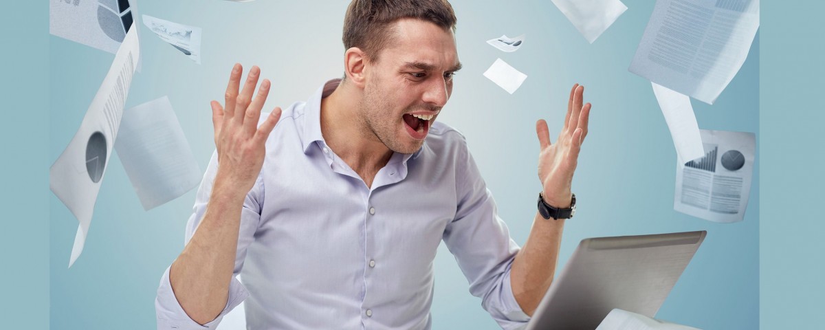 Man looking at laptop computer in frustration. Paper reports are falling nearby.