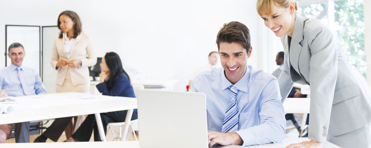 Group of office staff, focus is on a man on a laptop with a smiling woman looking at his screen