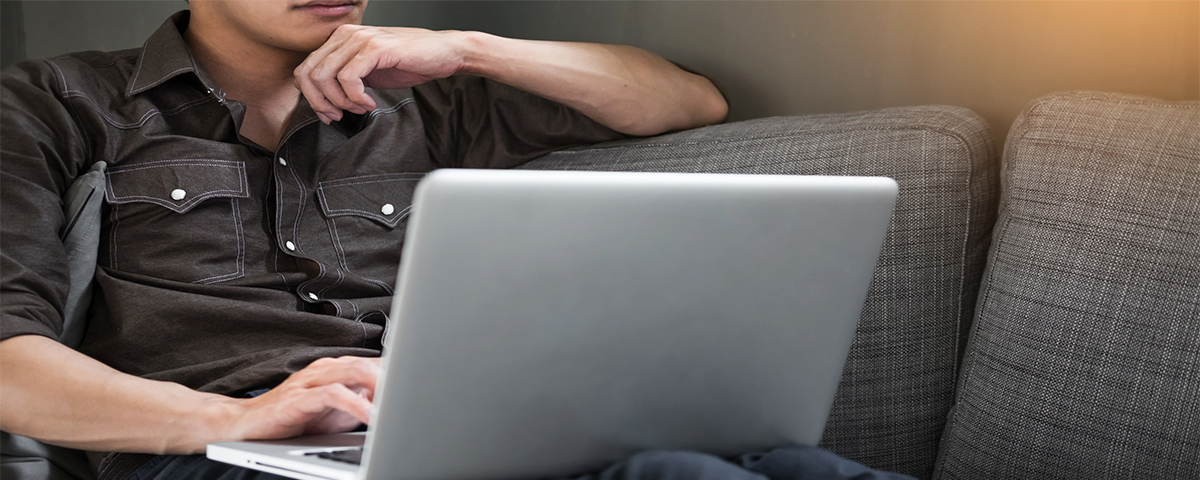 Man sitting on his couch. He is using a laptop and is clearly thinking deeply.