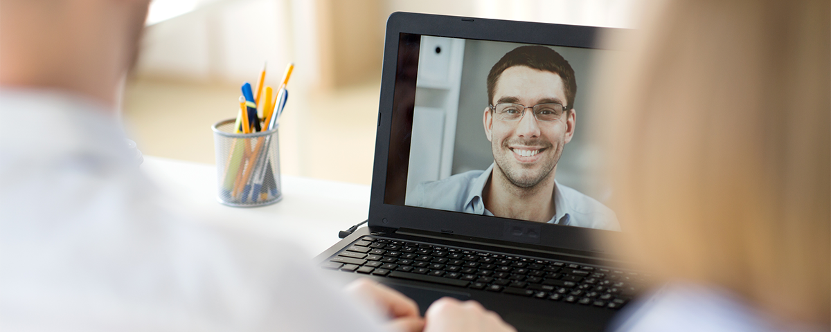 Image: Three people having a video conference together. Two are together in the same room, the other is a smiling man seen on a laptop screen.