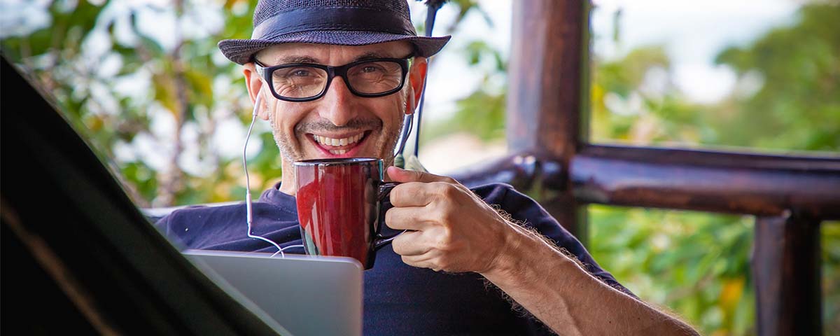 A smiling man sits in a hammock, drinking coffee while working remotely from his laptop