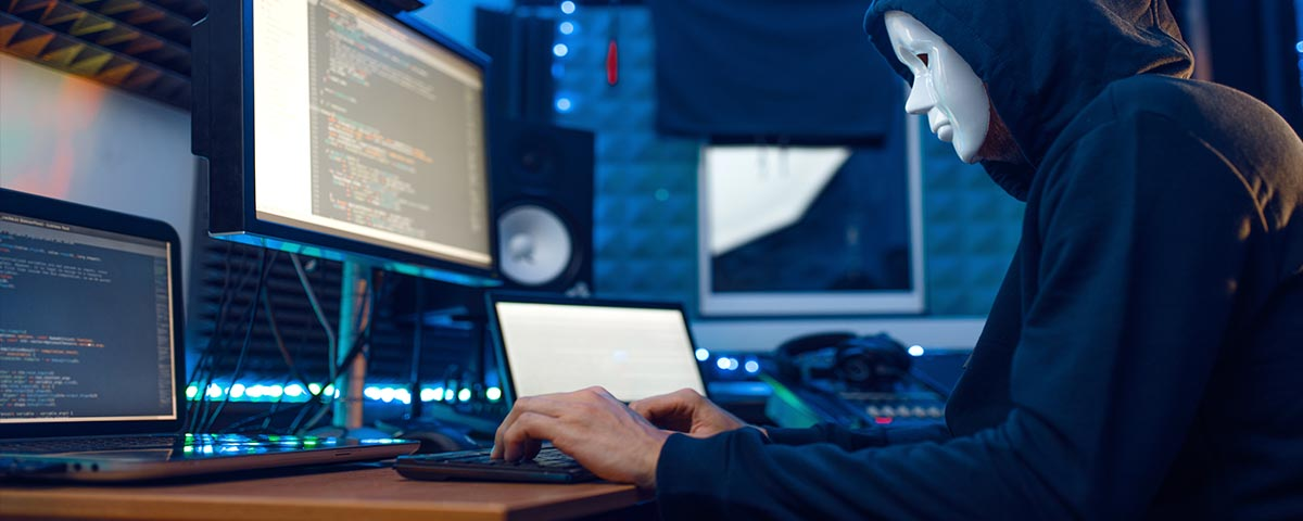 Person wearing a hooded shirt and a white mask, sitting in front of multiple computer monitors