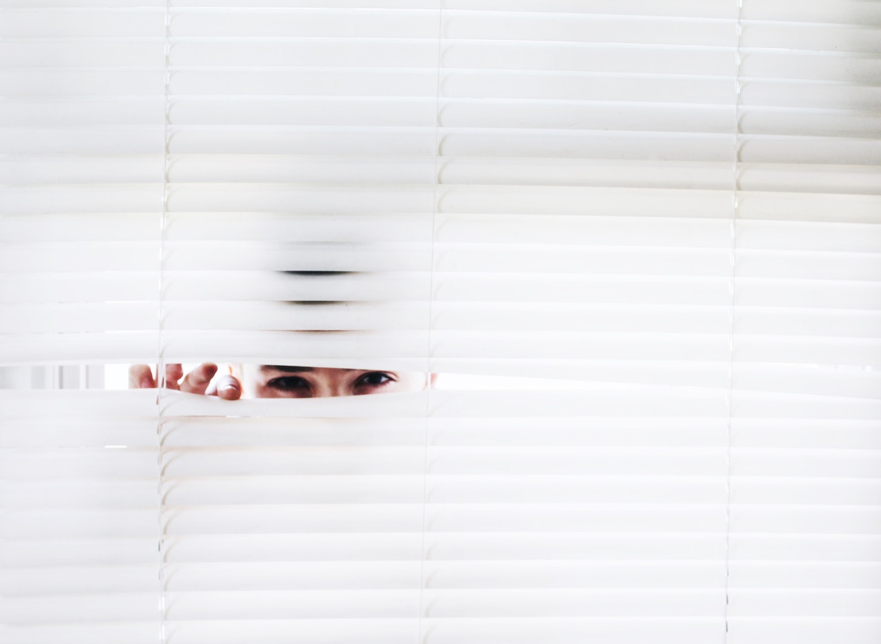 A barely visible man peeks through white blinds. Only his eyes and fingers are visible behind the blinds.