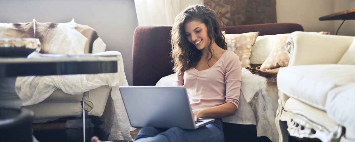 A woman working from home on her laptop, sitting on the floor and smiling with a laptop in her lap.